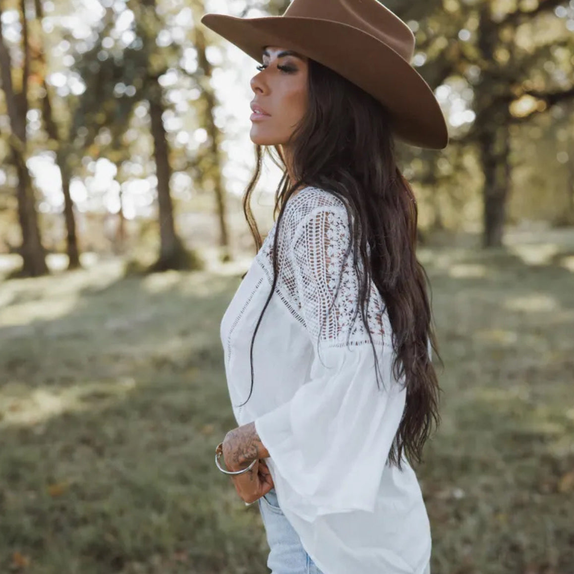 Woman wearing a white lace top and brown cowboy hat in a forest setting