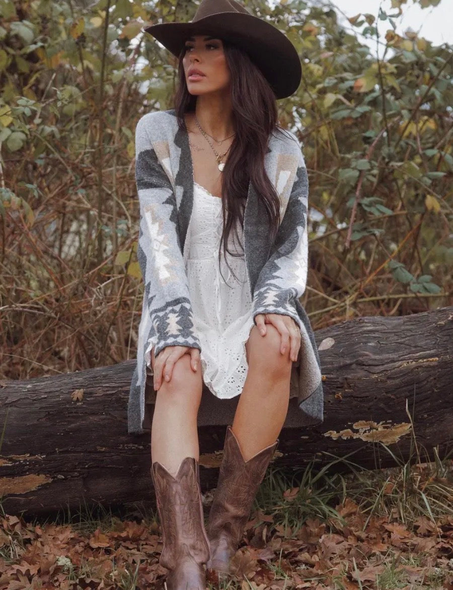 Woman in a patterned cardigan, white dress, and brown boots sitting on a log in a natural setting.