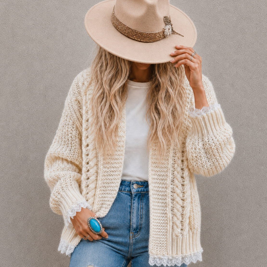 A woman wearing a cream chunky knit cardigan, a beige wide-brim fedora hat with a glittery band, a white t-shirt, and blue jeans, posing against a neutral gray wall.