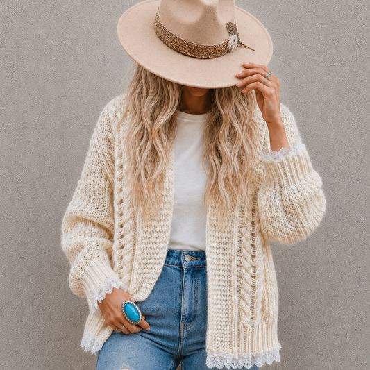 A woman wearing a cream chunky knit cardigan, a beige wide-brim fedora hat with a glittery band, a white t-shirt, and blue jeans, posing against a neutral gray wall.