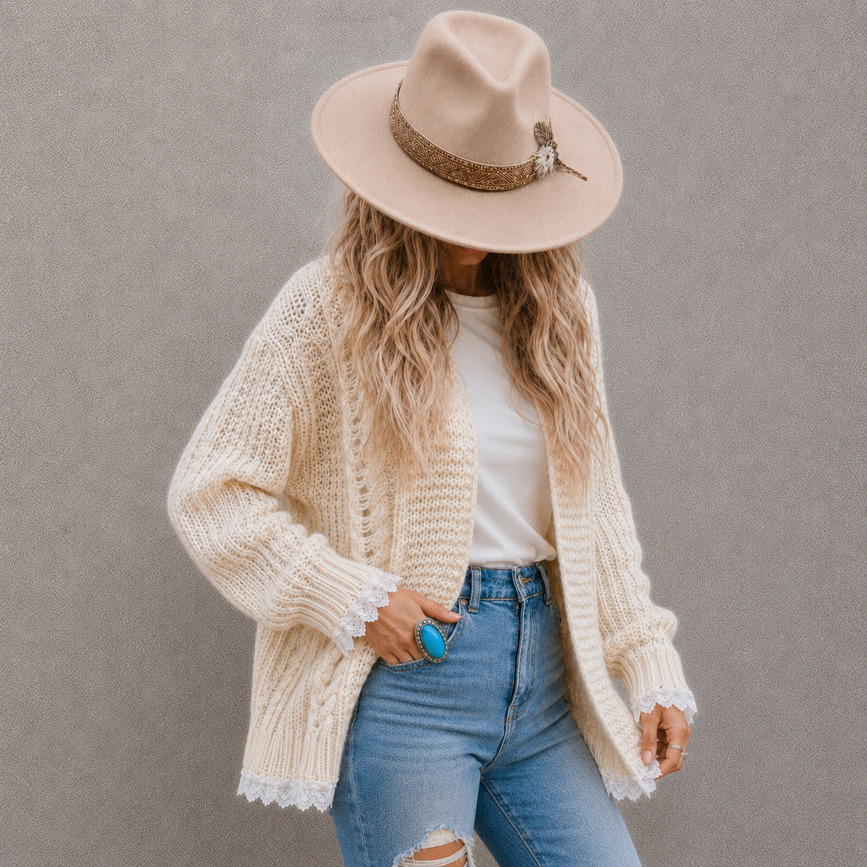 A woman wearing a cream-colored chunky knit cardigan, a beige wide-brim hat with a brown band, a white t-shirt, and light blue ripped jeans, standing against a textured gray wall.