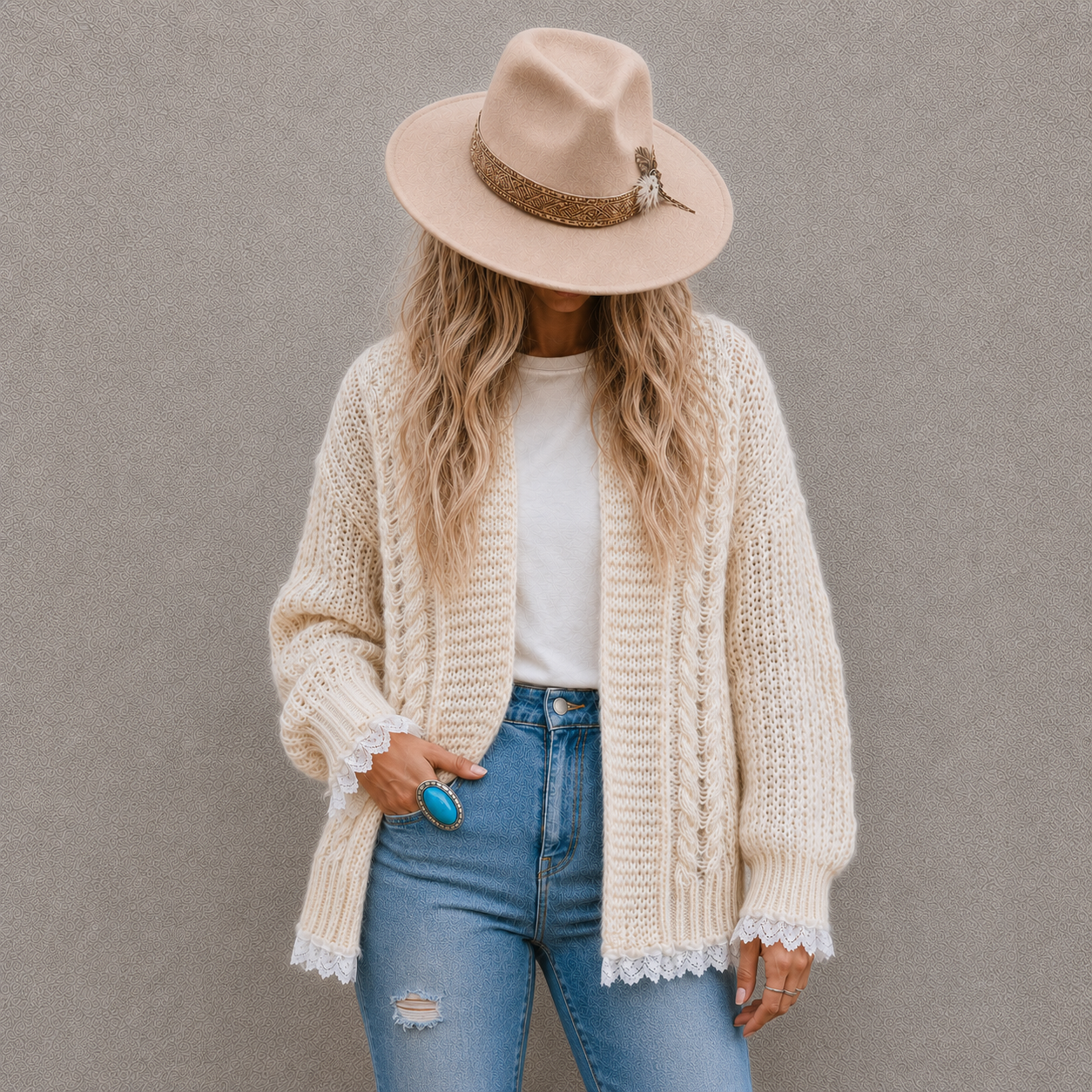 A woman wearing a cream cable-knit cardigan, a beige wide-brim hat, a white t-shirt, and blue jeans, standing against a textured gray wall.