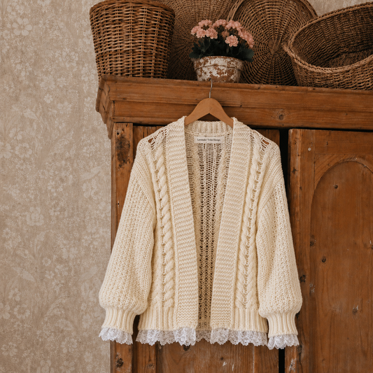 Cream cable-knit cardigan sweater hanging on a wooden hanger against a rustic wooden cabinet, with woven baskets and a small floral arrangement in the background.