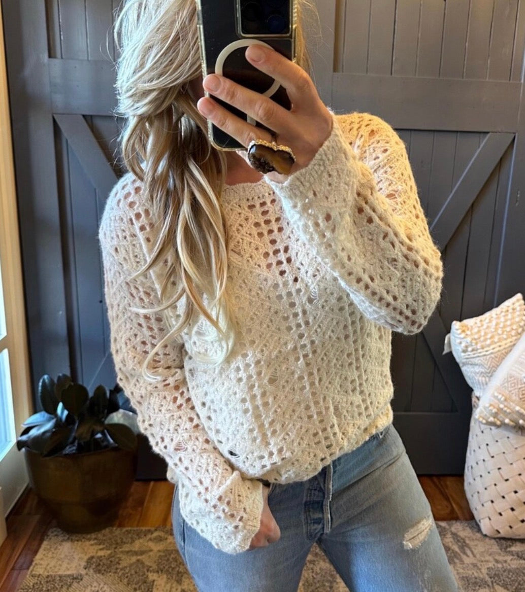 Person wearing a white crochet sweater and jeans, standing in a room with wooden flooring and a decorative pillow.