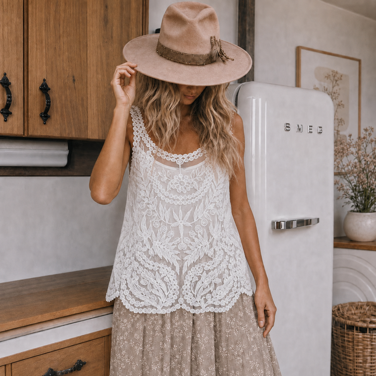 A woman wearing a beige wide-brim fedora hat and a white floral lace sleeveless top with a beige patterned skirt, standing in a rustic kitchen next to a white retro-style refrigerator.