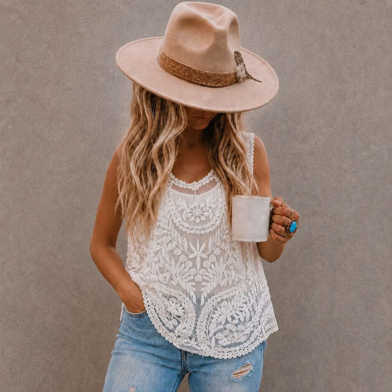 A woman wearing a beige wide-brim fedora hat, a white lace sleeveless top, and light blue jeans, holding a white mug against a textured gray wall.