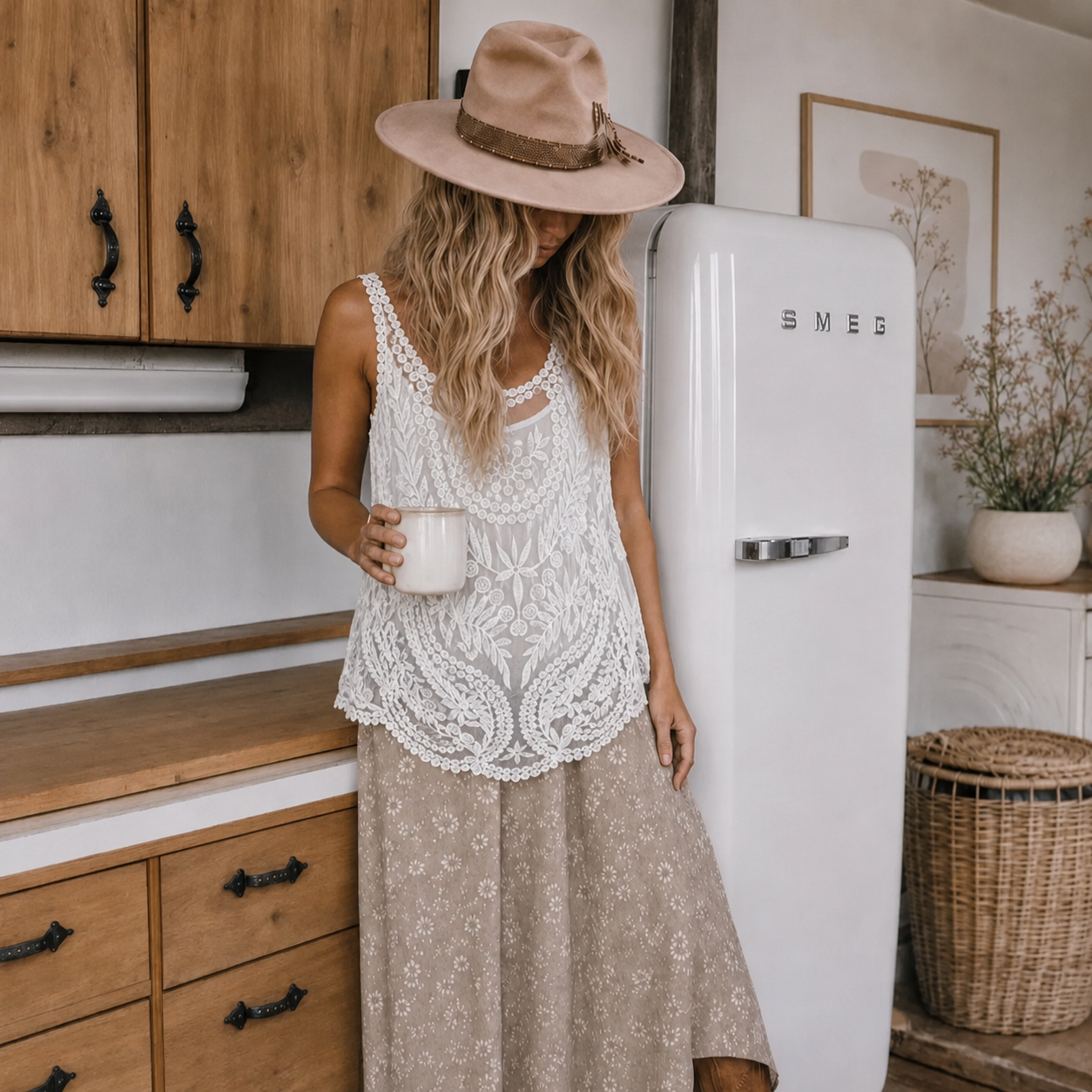 A woman stands in a rustic kitchen wearing a white crochet lace sleeveless top and a beige maxi skirt, with a wide-brimmed beige hat. She is holding a white mug next to a vintage white SMEG refrigerator.