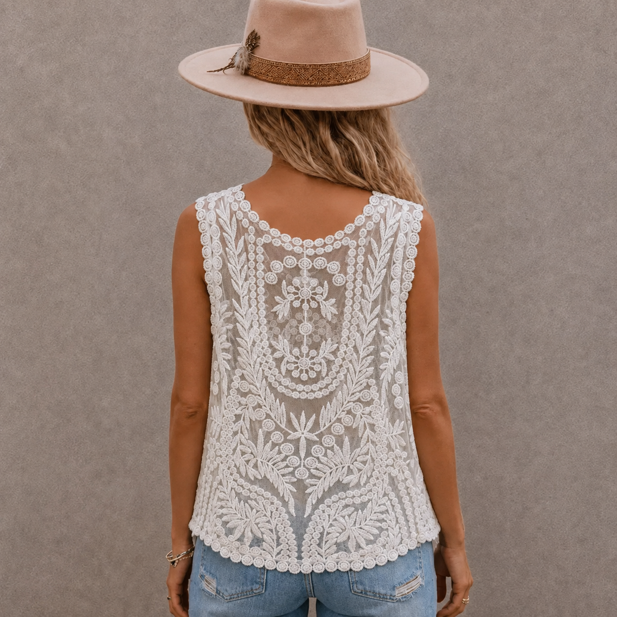 A woman viewed from behind wearing a sleeveless white lace top with intricate floral embroidery, paired with light blue jeans and a beige wide-brim hat against a textured gray wall.