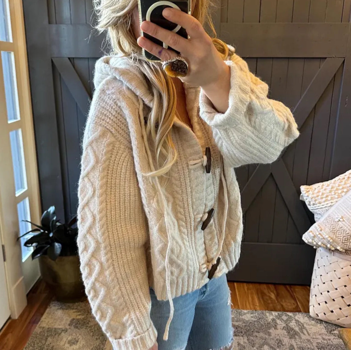 Person wearing a beige knitted cardigan taking a mirror selfie in a home setting.