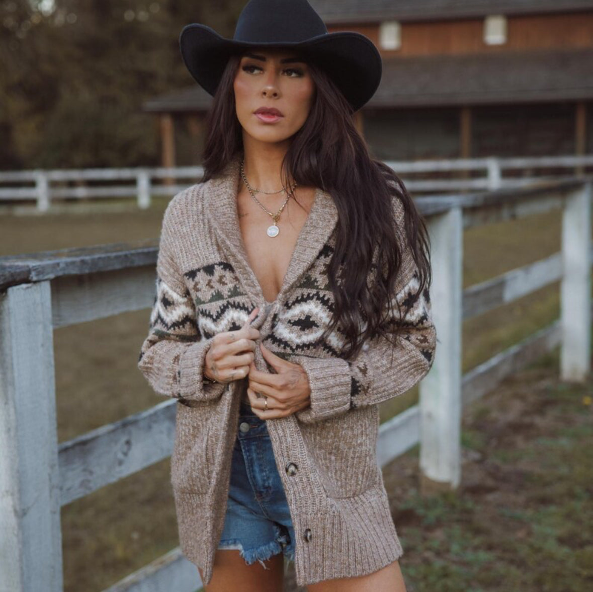 Woman wearing a patterned cardigan and black hat in an outdoor setting with a wooden fence and grass.