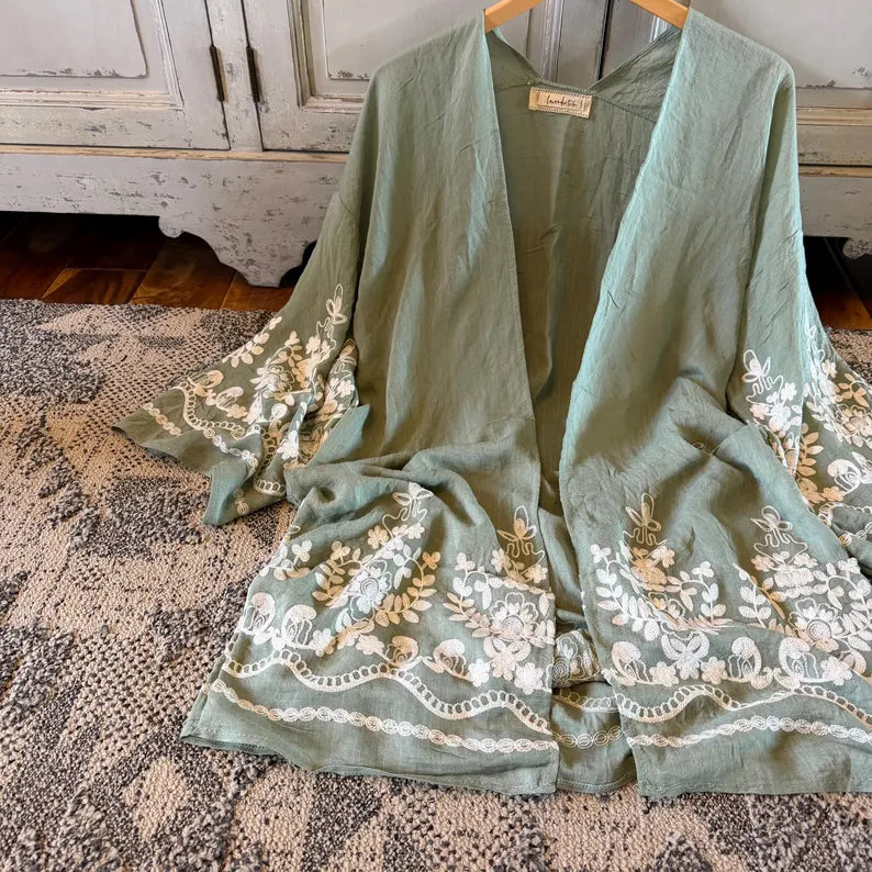 A sage green open-front kimono-style cardigan with white and gold floral embroidery, hanging on a wooden hanger on a patterned rug in front of a distressed white cabinet.