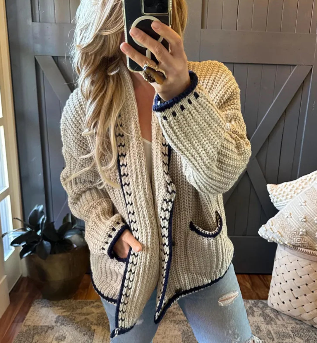Person wearing a beige knitted cardigan with black trim, taking a mirror selfie.