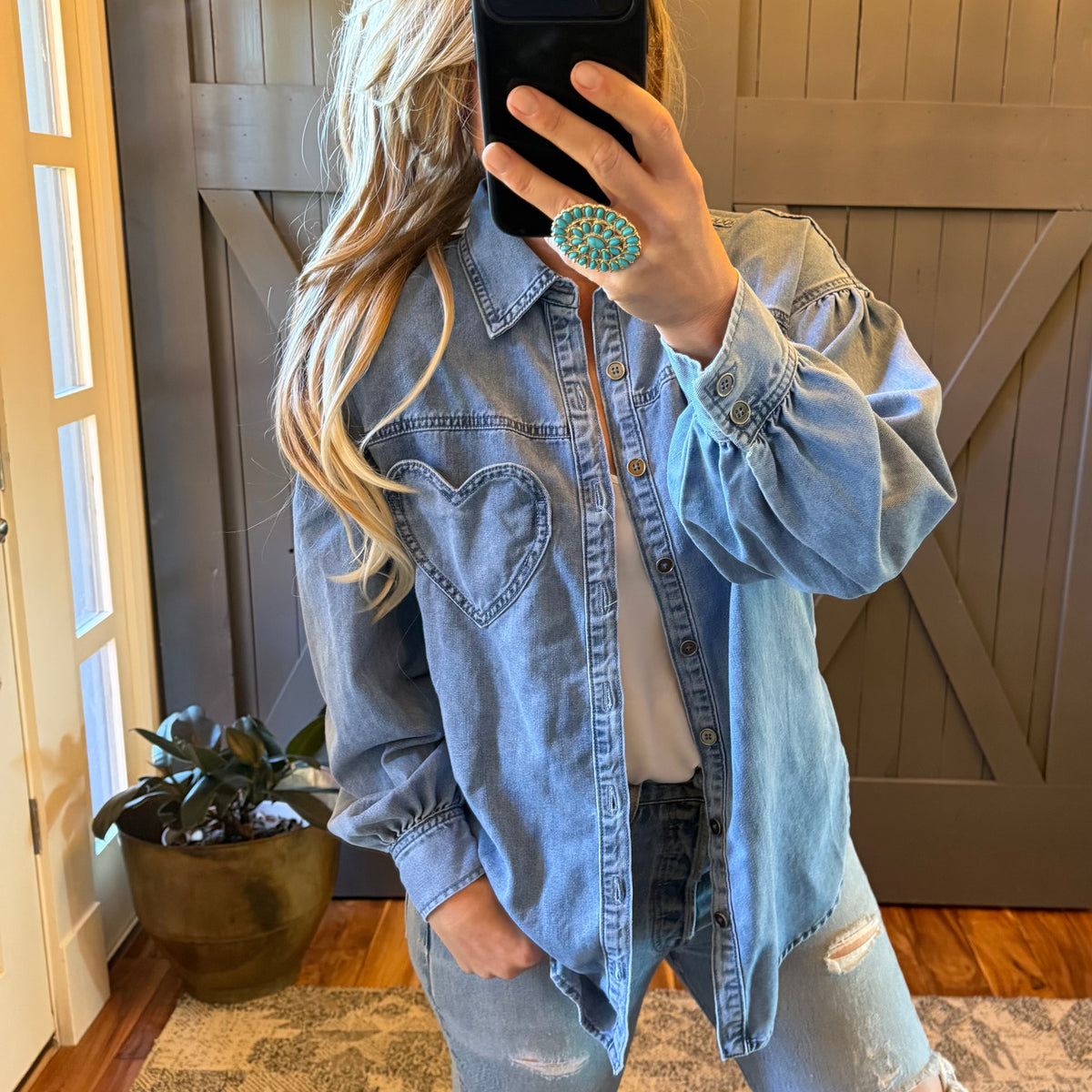 A woman takes a mirror selfie wearing an oversized light-wash blue denim shirt with a heart-shaped chest pocket, paired with distressed jeans and a white top. She is holding a black smartphone in front of her face, which is obscured by a turquoise patterned ring.