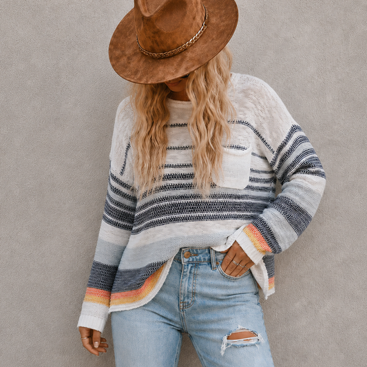 A woman wearing a brown wide-brim hat, a cream and navy striped knit sweater with orange stripes, and light blue ripped jeans, standing against a textured gray wall.