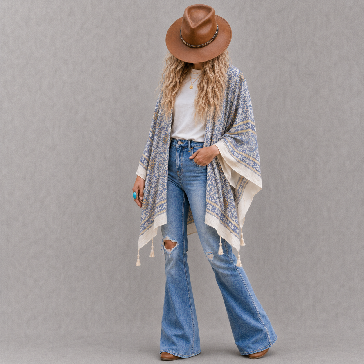 A woman stands against a textured gray studio background wearing a brown wide-brim hat, a white top, blue distressed flare jeans, and a light blue and white patterned poncho with tassels.