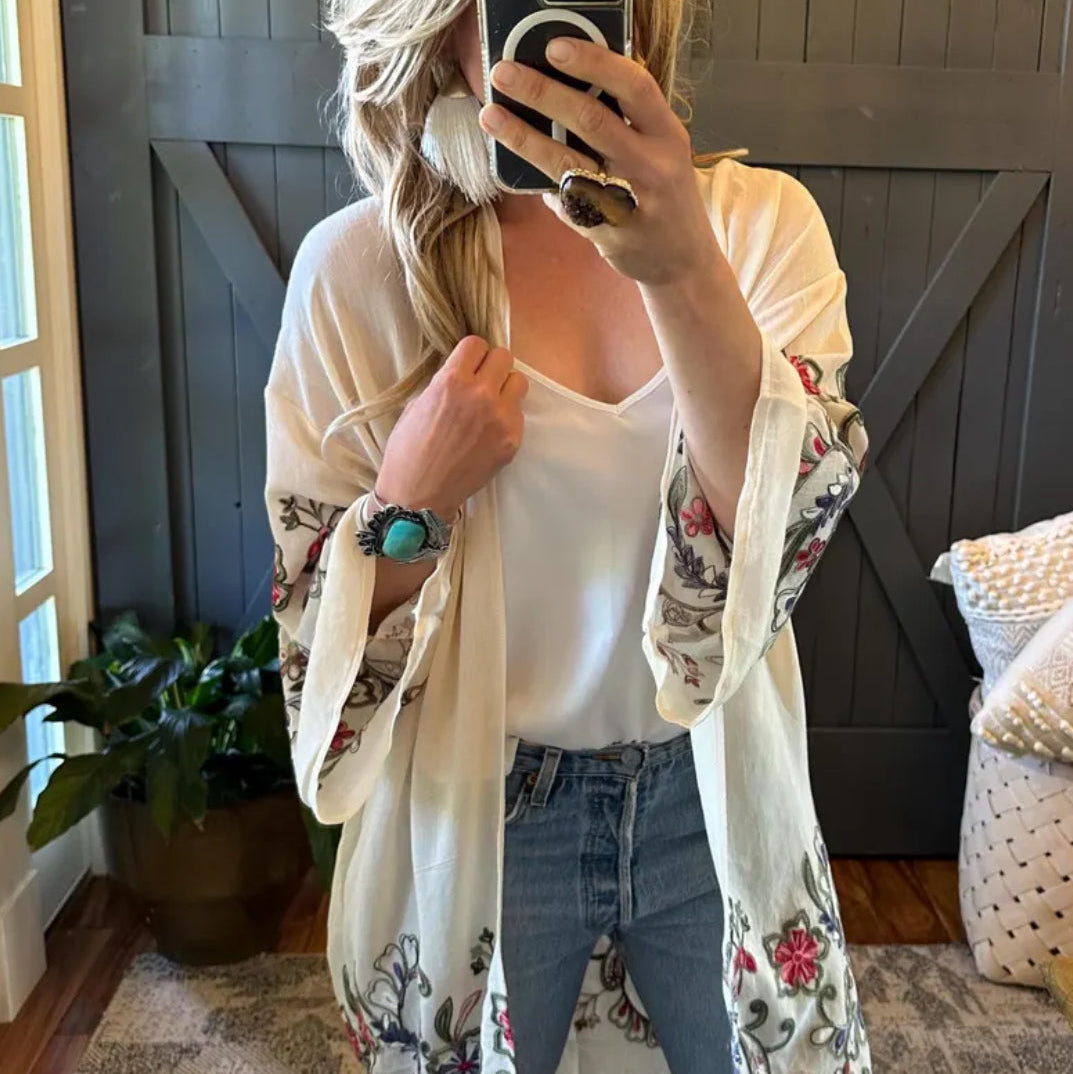 Person taking a mirror selfie wearing a floral kimono over a white top and jeans, with a home interior background.