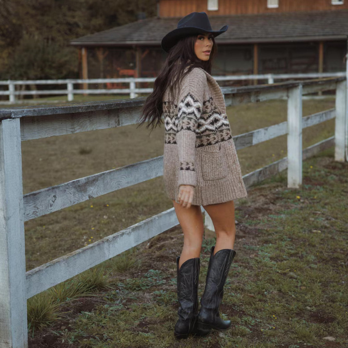 Woman in a patterned cardigan and cowboy boots standing near a wooden fence.