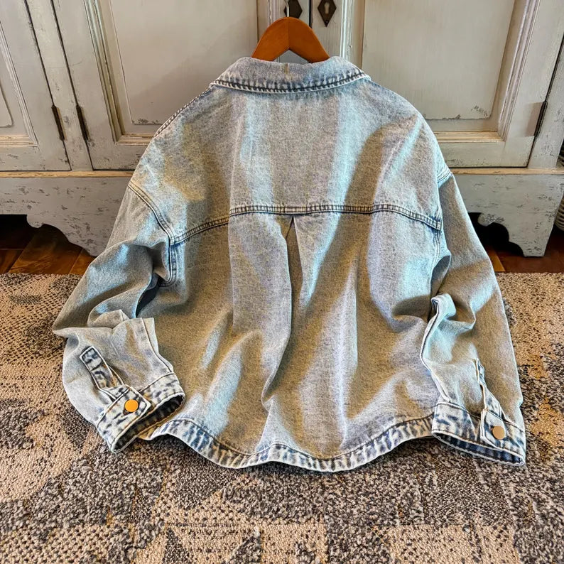 A light-wash blue denim jacket hanging on a wooden hanger, showing the back view with a central seam and buttoned cuffs, set against a rustic white cabinet and patterned rug.