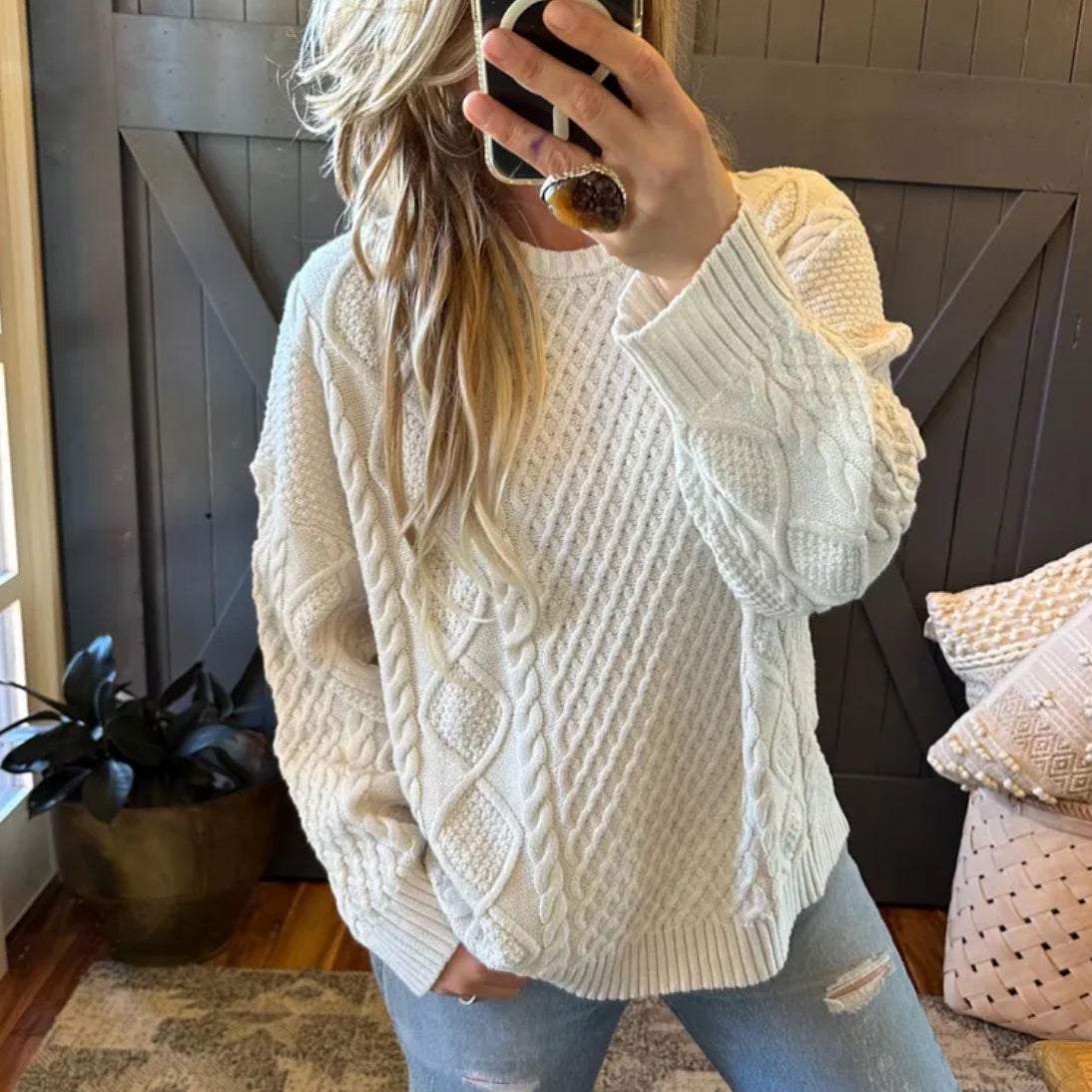 Person wearing a cream sweater taking a mirror selfie in a home setting.