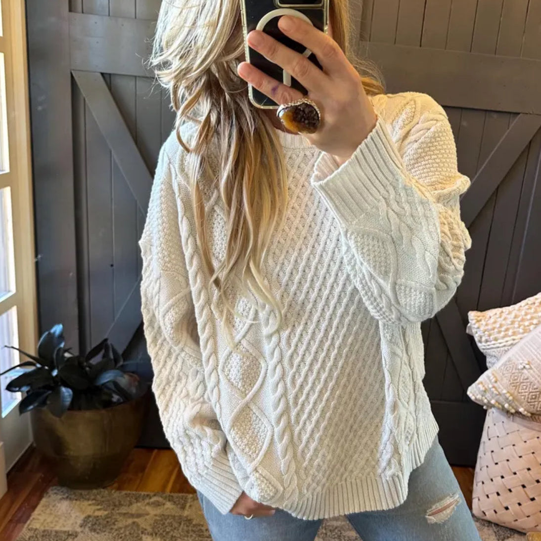 Person wearing a white cable knit sweater taking a mirror selfie in a home setting.