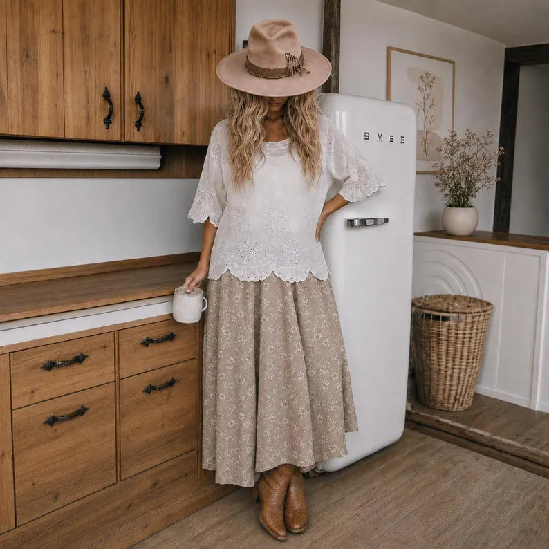 A woman stands in a rustic kitchen wearing a bohemian-style outfit featuring a white lace short-sleeve top and a beige floral maxi skirt, paired with a wide-brimmed straw hat and brown ankle boots. She is positioned next to a white vintage-style refrigerator.
