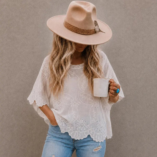 A woman wearing a white eyelet lace short-sleeve blouse with a scalloped hem, paired with light blue jeans and a beige wide-brim hat, holding a white mug against a textured gray wall.