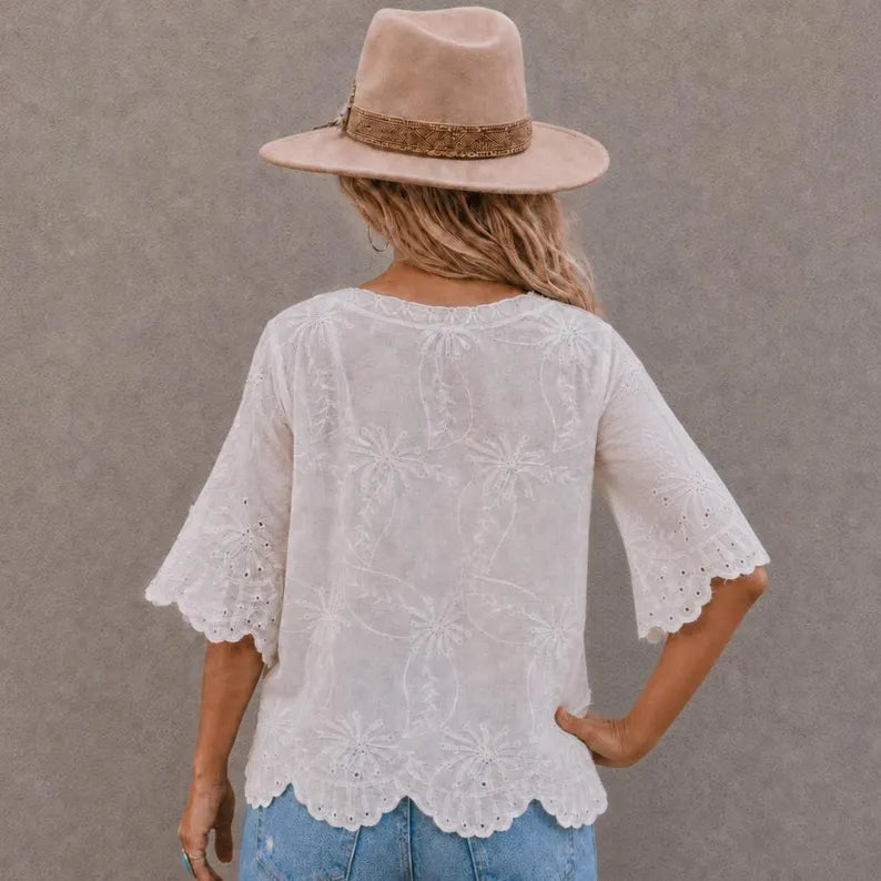 A woman shown from the back wearing a white eyelet lace short-sleeve blouse with a scalloped hem, paired with blue jeans and a beige wide-brim hat against a textured gray wall.