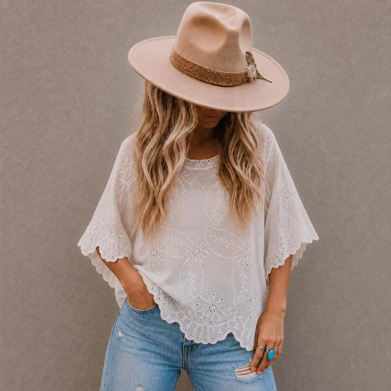 A woman wearing a cream eyelet lace short-sleeve top, blue jeans, and a beige wide-brim hat with a brown band, standing against a textured gray wall.