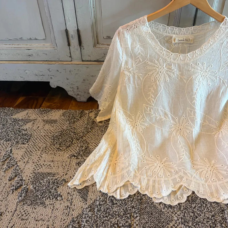 An off-white eyelet lace blouse with floral embroidery and a scalloped hem, hanging on a wooden hanger on a patterned rug in front of a distressed white cabinet.