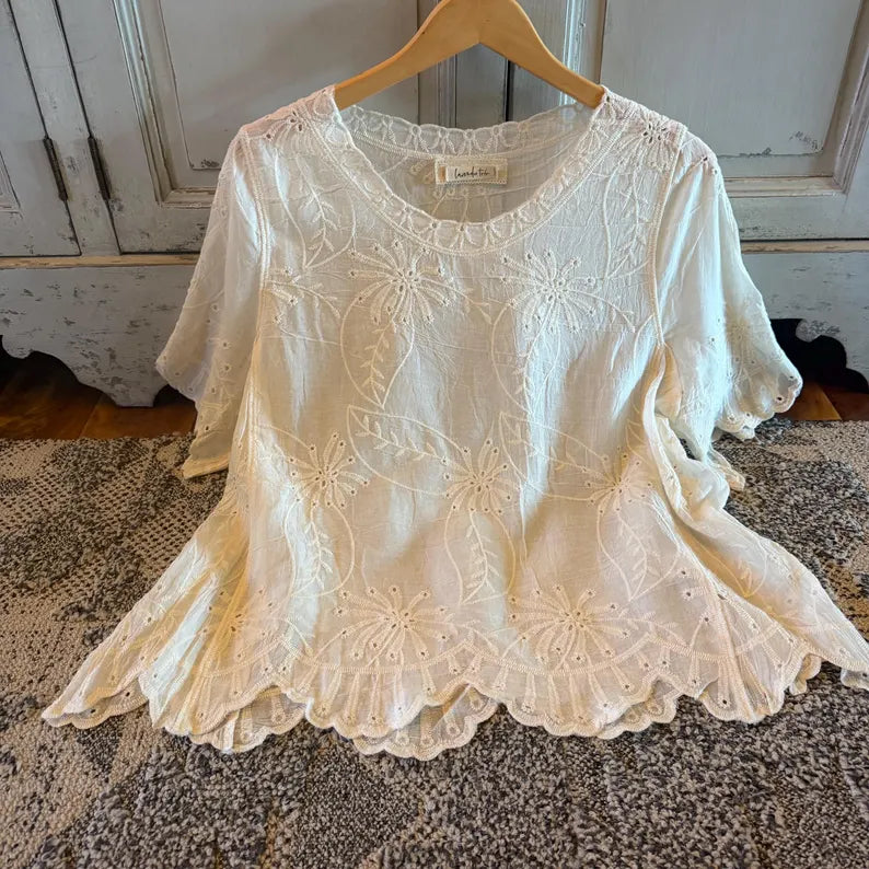 An off-white eyelet lace blouse with floral embroidery and a scalloped hem, hanging on a wooden hanger on a patterned rug in front of a rustic white cabinet.