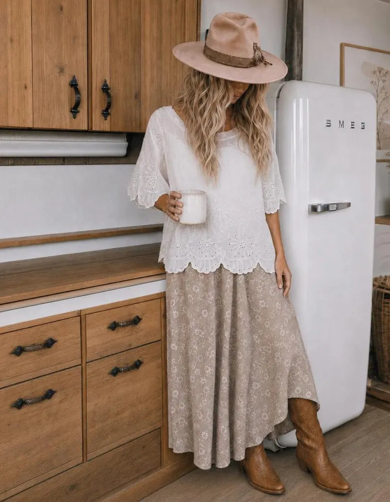 A woman stands in a rustic kitchen wearing a bohemian-style outfit consisting of a white lace short-sleeve top, a beige floral maxi skirt, a wide-brimmed beige hat, and brown boots. She is holding a white mug next to a white retro SMEG refrigerator.