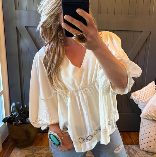 A woman takes a mirror selfie wearing a cream-colored puff-sleeve blouse with a V-neck and gathered waist, paired with light-wash distressed jeans. She is wearing a large statement ring and a turquoise bracelet, standing in front of a dark wood-paneled wall.