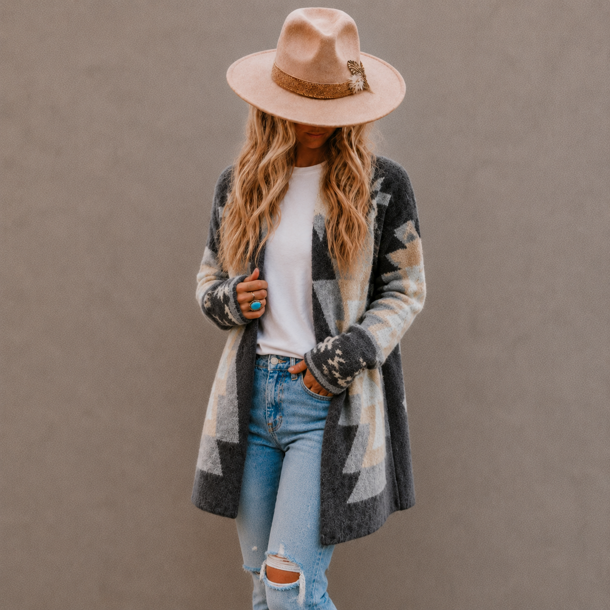 A woman wearing a beige wide-brim fedora hat, a gray, black, and beige geometric pattern knit cardigan over a white t-shirt and light-wash distressed jeans, standing against a solid gray background.