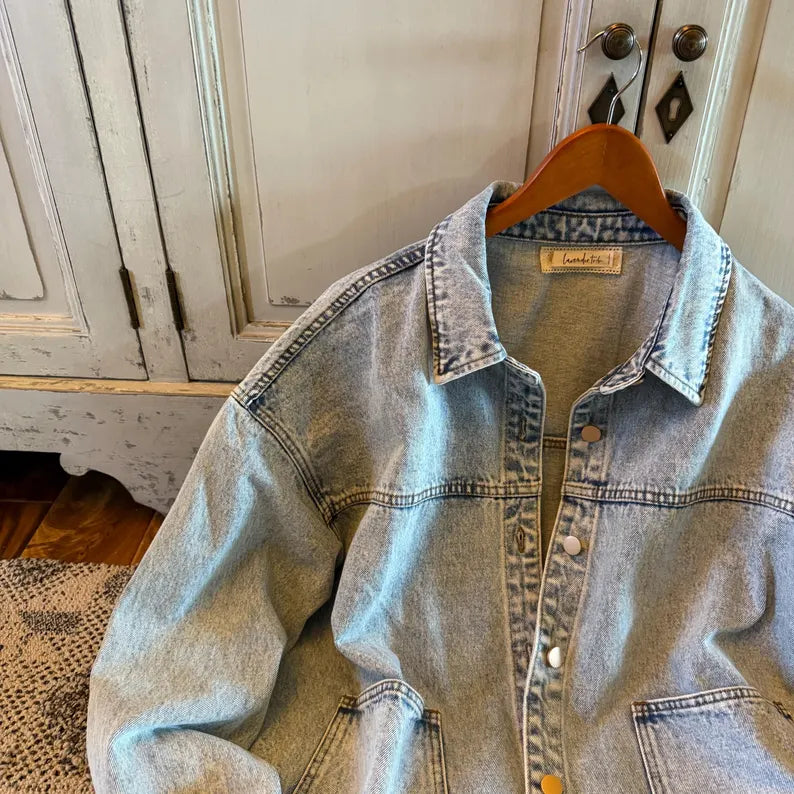 A light-wash denim button-up shirt jacket hanging on a wooden hanger against a distressed white cabinet background.