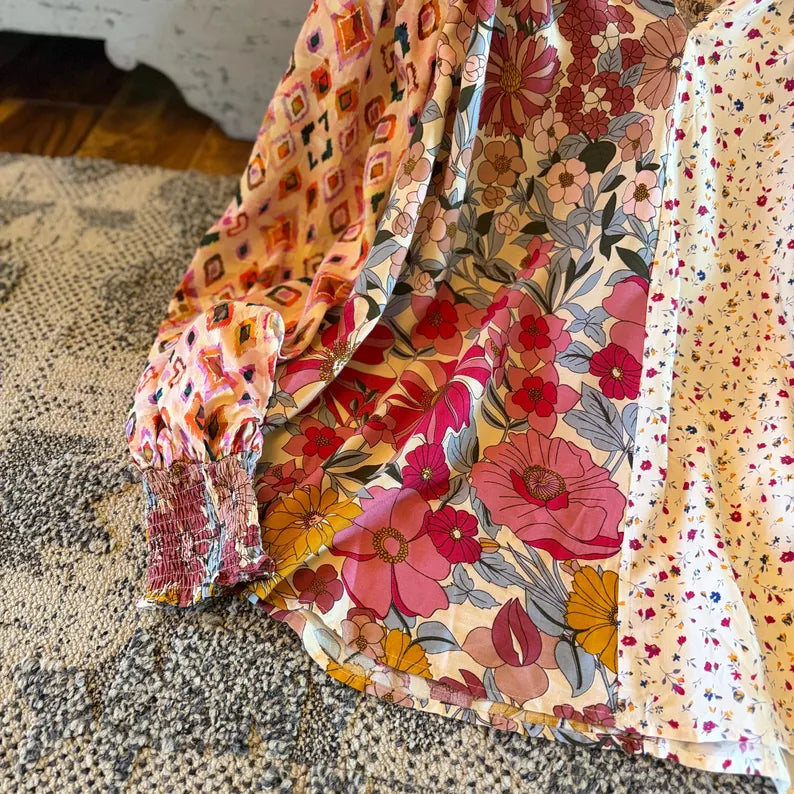 A close-up of a multicolor floral and patterned blouse draped on a textured rug. The fabric features a busy print with pink, red, yellow, and orange flowers, geometric shapes, and a small red floral dot pattern on a cream background.