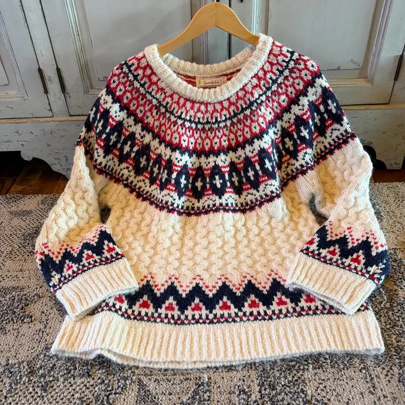 A chunky knit crewneck sweater with a cream base and red and navy blue Fair Isle patterns, hanging on a wooden hanger on a patterned rug in front of a rustic cabinet.