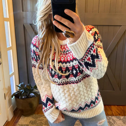 A woman takes a mirror selfie wearing a chunky knit crewneck sweater with a cream base and a colorful Fair Isle pattern in red, navy, and white. She is wearing light-wash distressed jeans and a large statement ring, standing in front of a gray wooden barn door.