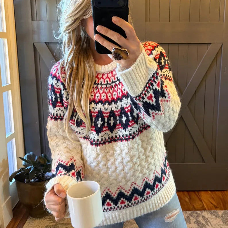 A woman takes a mirror selfie wearing a chunky knit crewneck sweater with a multicolor Fair Isle pattern in cream, red, and navy blue. She is holding a white ceramic mug in one hand and a black smartphone in the other, standing in front of a wooden barn-style door.