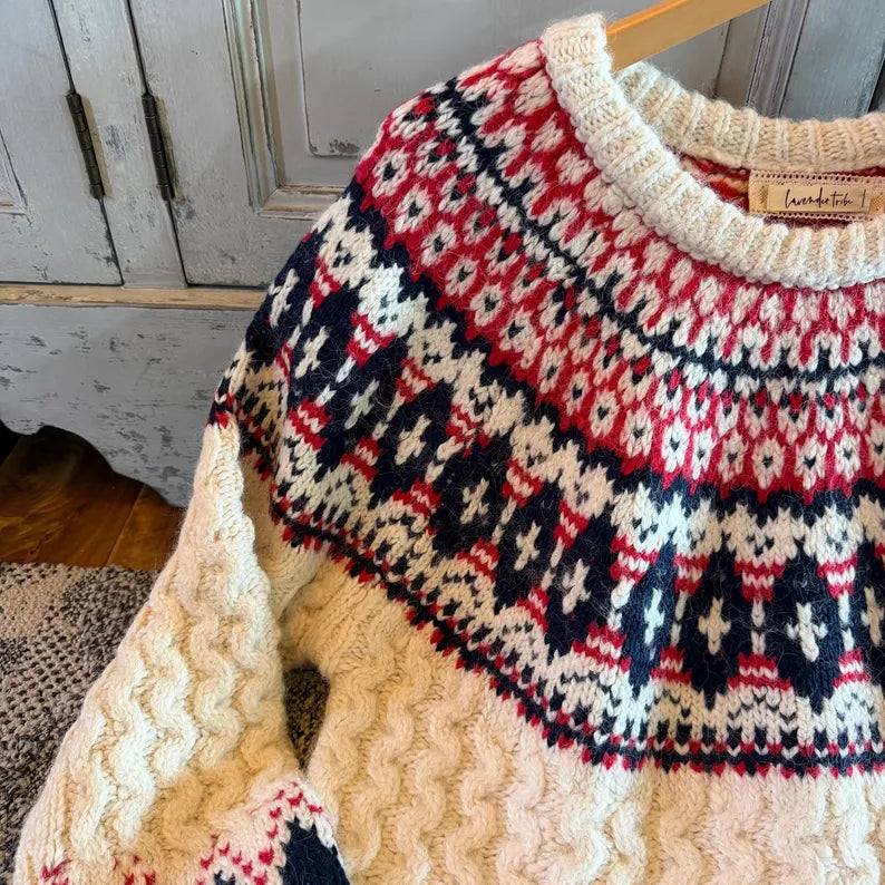 A close-up of a knitted sweater hanging on a wooden hanger, featuring a cream base with a red and navy blue Fair Isle pattern across the yoke and upper sleeves. The knit texture is chunky and cozy, and a brand label is visible inside the collar.