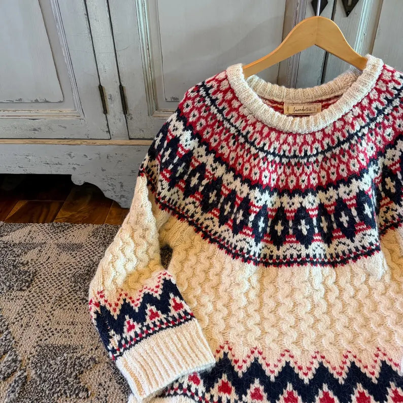 A cream-colored cable-knit sweater with red and navy blue Fair Isle patterns, hanging on a wooden hanger against a rustic white cabinet background.