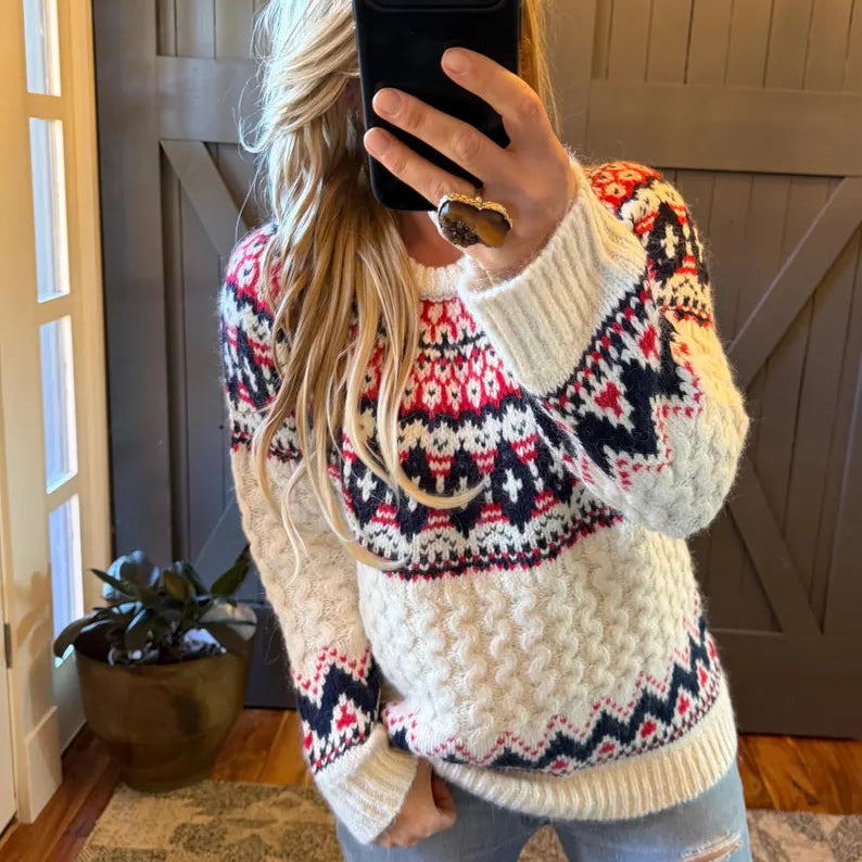 A woman takes a mirror selfie wearing a cream-colored knit sweater with a colorful Fair Isle pattern in red, navy, and white. She is wearing light-wash distressed jeans and holding a black smartphone to take the photo in a home setting.