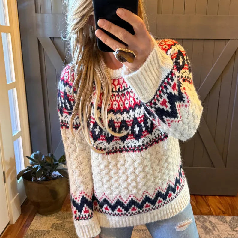 A woman takes a mirror selfie wearing a chunky knit crewneck sweater with a cream base and colorful Fair Isle patterns in red, pink, and navy blue. She is wearing light-wash distressed jeans and a gold necklace, standing in front of a dark wood barn-style door.