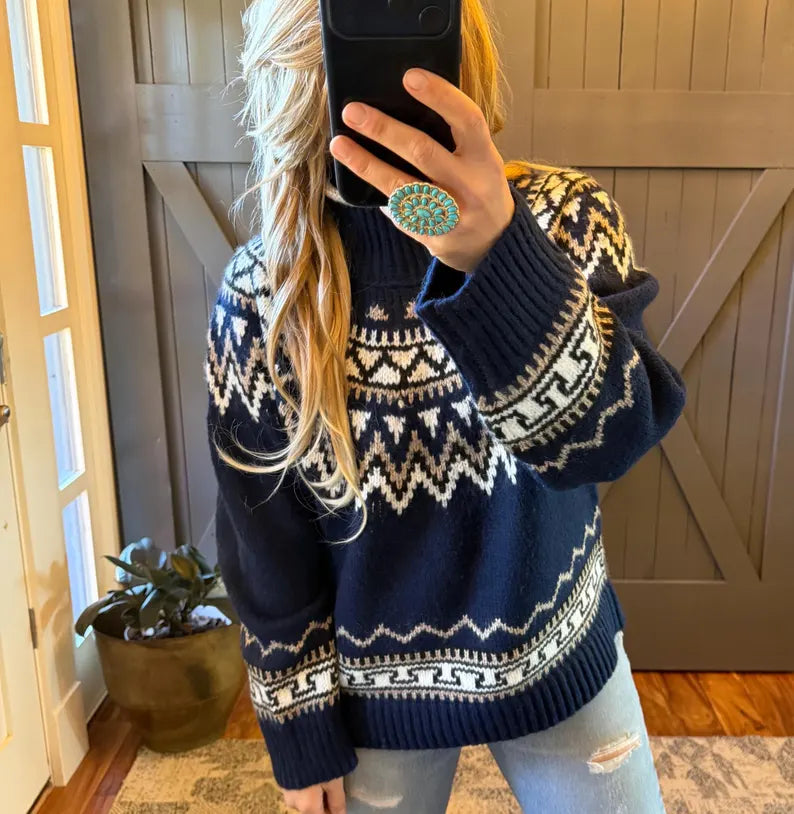 A woman takes a mirror selfie wearing a navy blue knit sweater with a white Fair Isle pattern, paired with light blue jeans. She is holding a black smartphone to take the photo, with a wooden barn door and a potted plant in the background.