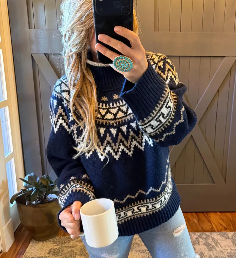A woman takes a mirror selfie wearing a navy blue knit sweater with a cream geometric Fair Isle pattern. She is holding a white ceramic mug and standing in front of a gray wooden barn door.