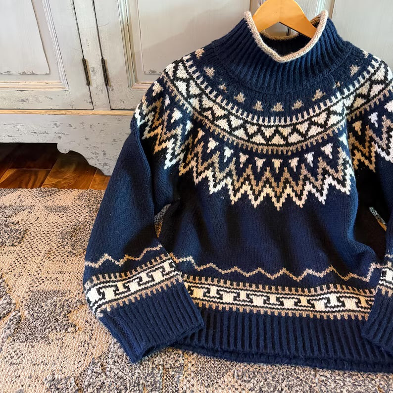 A navy blue knit sweater with a cream and beige Fair Isle pattern, hanging on a wooden hanger on a patterned rug in front of a rustic white cabinet.
