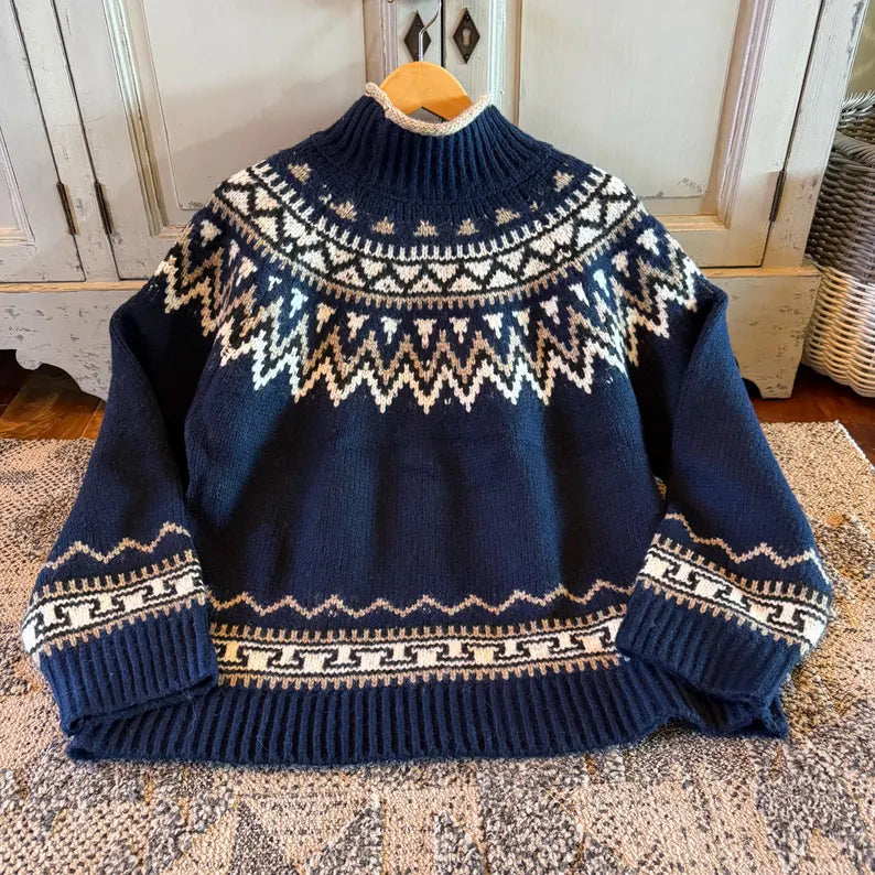 A navy blue knit sweater with cream-colored Fair Isle patterns, displayed on a wooden hanger over a patterned rug in front of a rustic white cabinet.