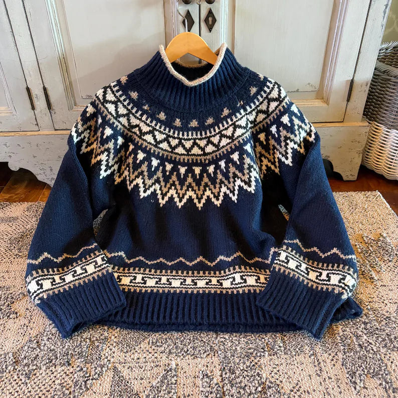 A navy blue knit sweater with a cream and white Fair Isle pattern, hanging on a wooden hanger on a patterned rug in front of a rustic white cabinet.