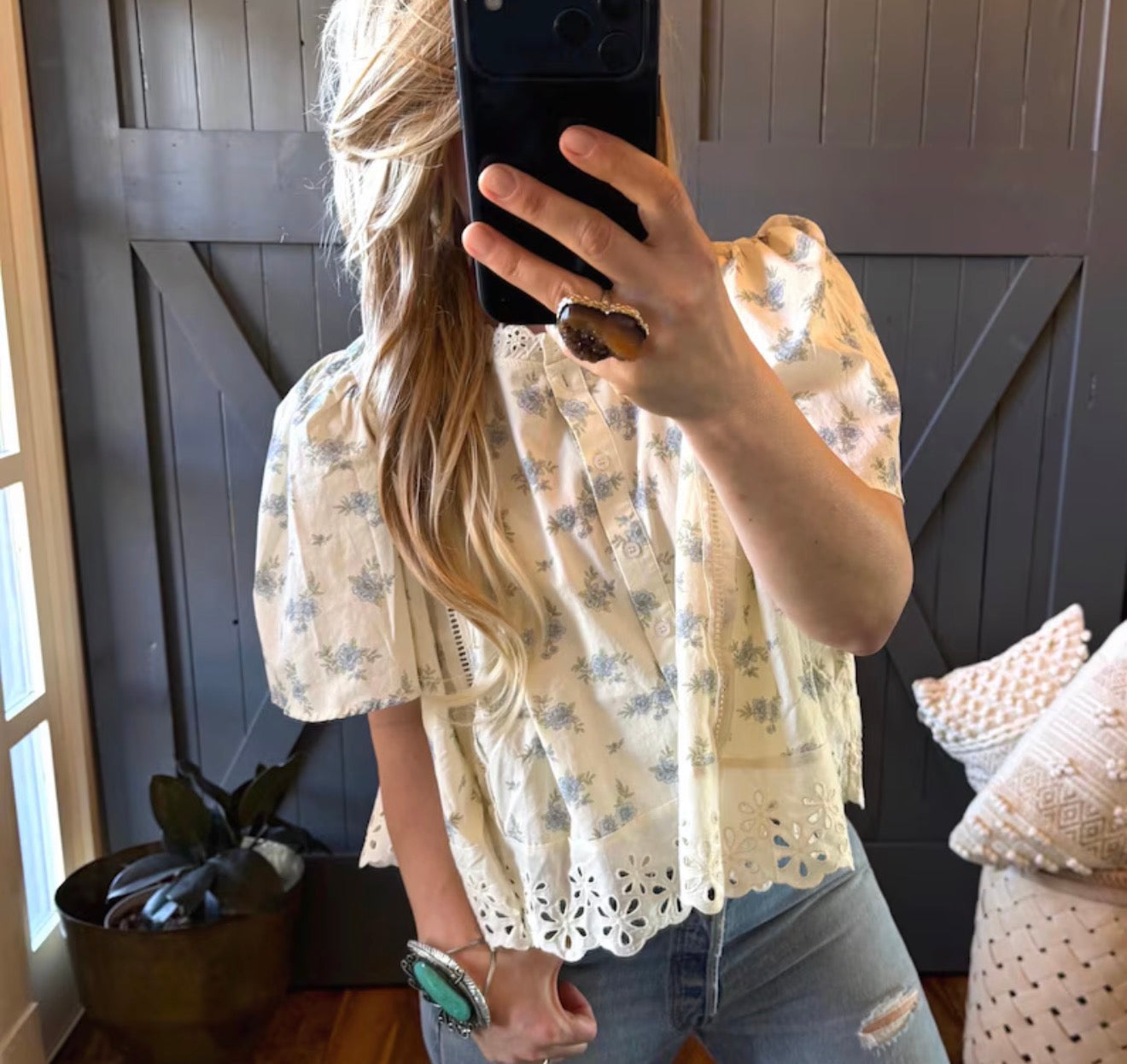 Person wearing a floral blouse taking a mirror selfie in a room with a dark wall and plant.