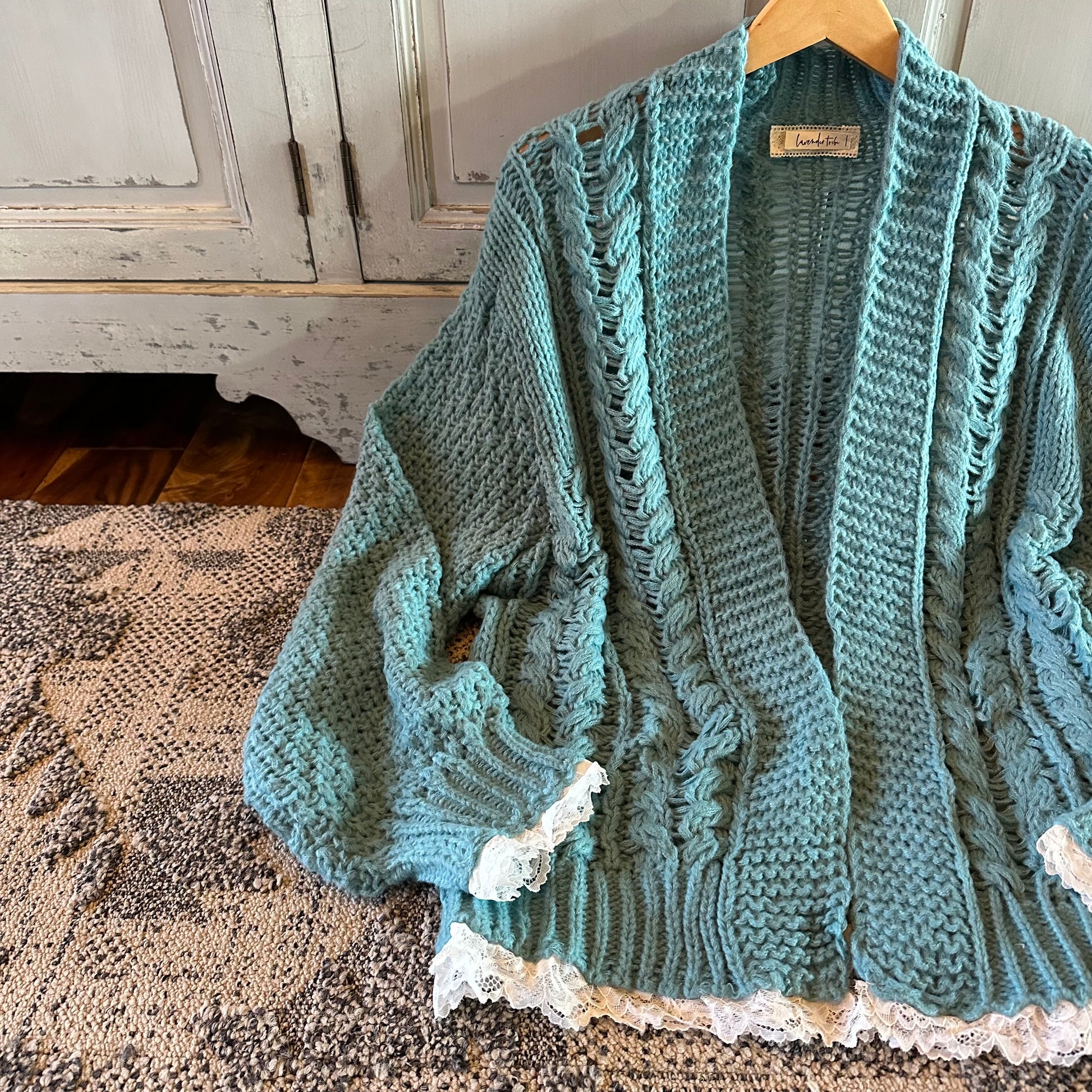 Knitted cardigan with lace trim on a wooden hanger against a rustic wooden cabinet background.