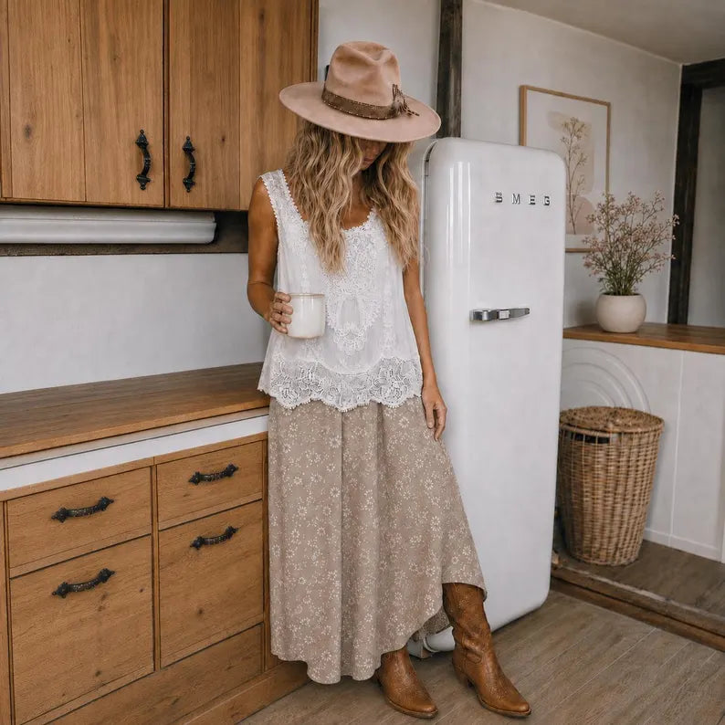 A woman stands in a rustic kitchen wearing a bohemian-style outfit consisting of a white lace sleeveless top, a beige floral maxi skirt, a wide-brimmed tan hat, and brown leather boots. She is positioned in front of a vintage white SMEG refrigerator.